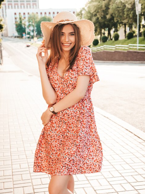 Portrait of young beautiful smiling hipster girl in trendy summer sundress.Sexy carefree woman posing on the street background in hat at sunset. Positive model outdoors