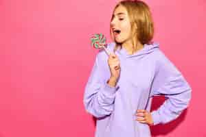 Free photo portrait of young beautiful smiling hipster woman in trendy summer hoodie. sexy carefree woman posing near pink wall. positive model with lollipop