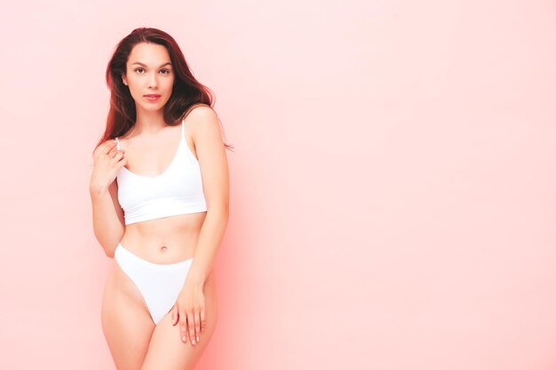 Portrait of young beautiful smiling woman in white lingerie. Sexy carefree cheerful model in underwear posing near pink wall in studio. Positive and happy female enjoying morning