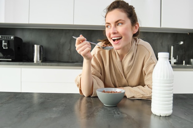 Free photo portrait of young beautiful woman in bathrobe eating cereals for breakfast leans on kitchen worktop