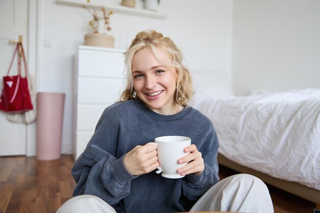 Portrait of young beautiful woman in casual clothes sitting on bedroom floor with cup of tea