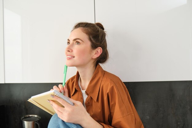 Portrait of young beautiful woman in the kitchen looking aside and thinking holding planner writing
