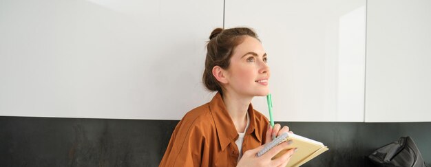 Portrait of young beautiful woman in the kitchen looking aside and thinking holding planner writing