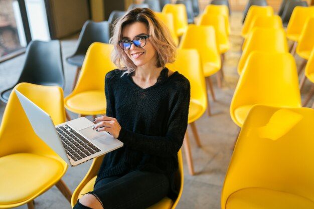 Portrait of young beautiful woman sitting in lecture hall, working on laptop, wearing glasses, classroom