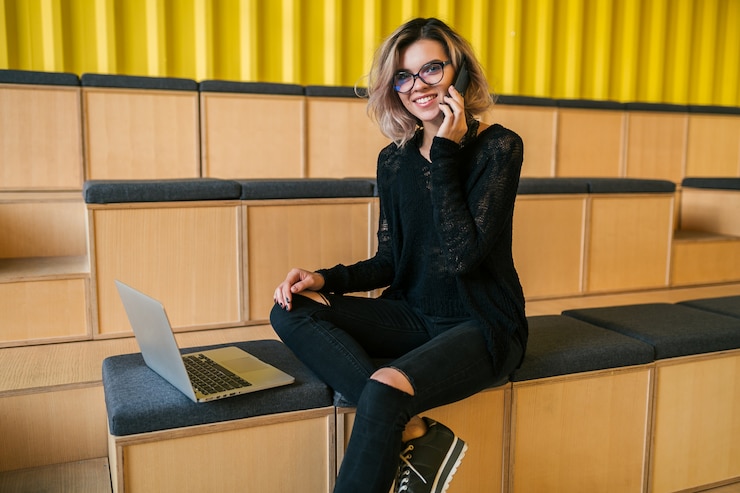 Free Photo portrait of young beautiful woman sitting in lecture hall, working on laptop, wearing glasses, modern auditorium, student education online, freelancer, smiling, talking on phone