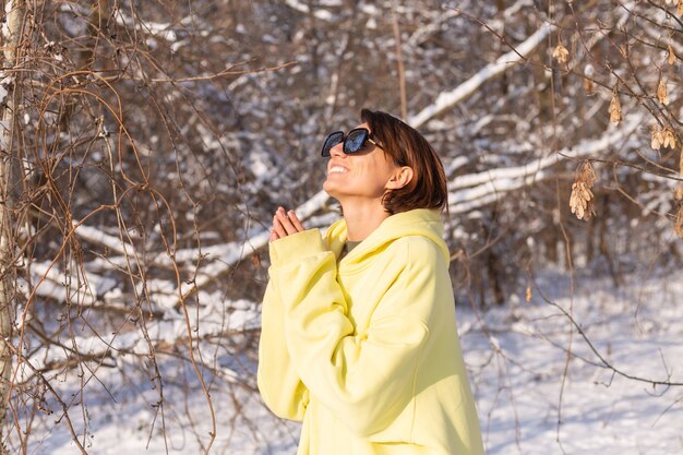 晴れた日の雪景色の冬の森で、サングラスをかけ、太陽と雪を楽しんで、黄色の大きなプルオーバーに身を包んだ若い美しい女性の肖像画