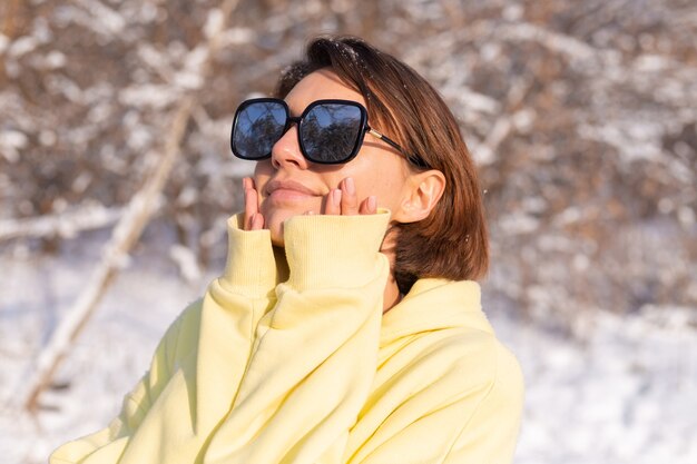 晴れた日の雪景色の冬の森で、サングラスをかけ、太陽と雪を楽しんで、黄色の大きなプルオーバーに身を包んだ若い美しい女性の肖像画