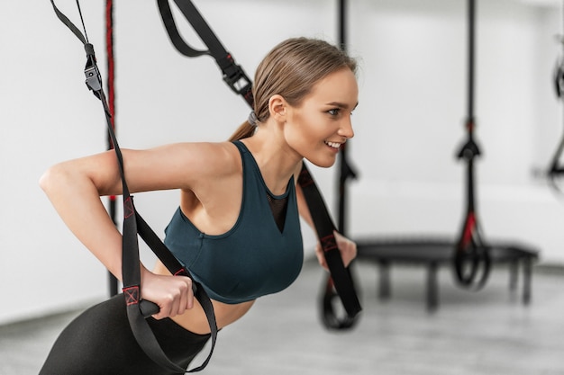 Portrait of young beautiful woman in sportswear training arms with trx fitness straps in the gym doing push ups