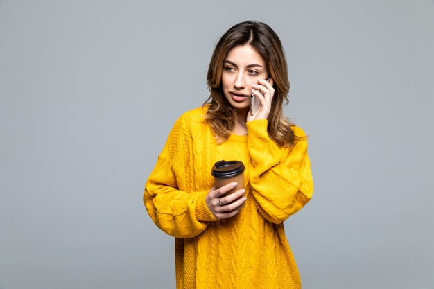 Portrait of young beautiful woman in yellow top, holding cardboard cup of takeaway coffee, smiling happily isolated on grey wall