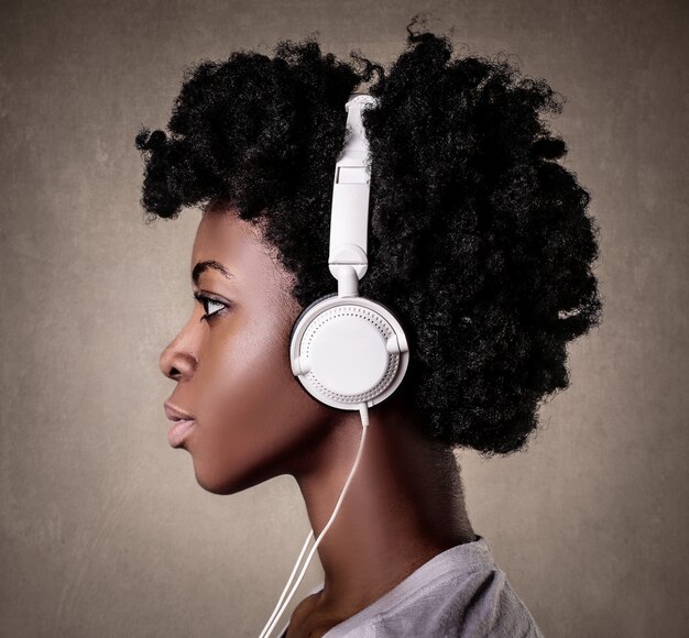Портрет молодой черной женщины с вьющимися волосами, слушающей музыку в белых наушниках