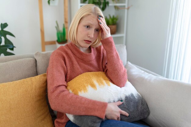 Портрет молодой блондинки, сидящей дома на диване с головной болью и мигренью. Красивая женщина, страдающая от хронических ежедневных головных болей.