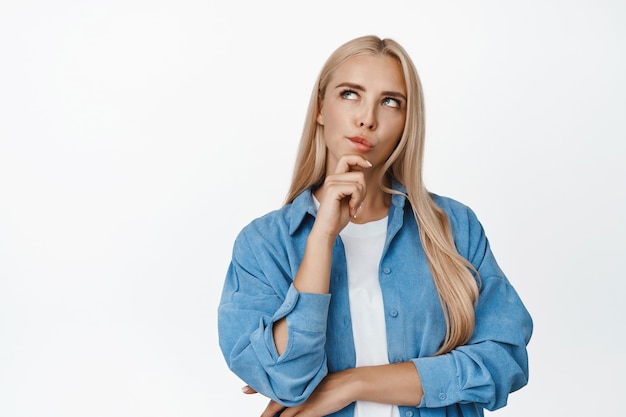 Portrait of young blond woman thinking, looking serious at upper left corner thoughtful, making decision, standing on white