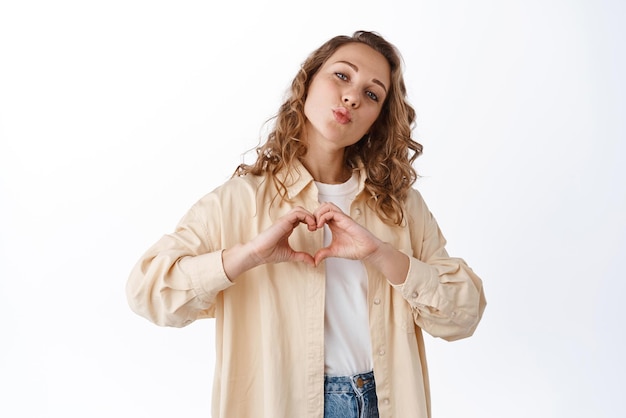 Portrait of young blond woman with curly hair showing love heart gesture pucker lips say I love you express admiration making kissing face white background