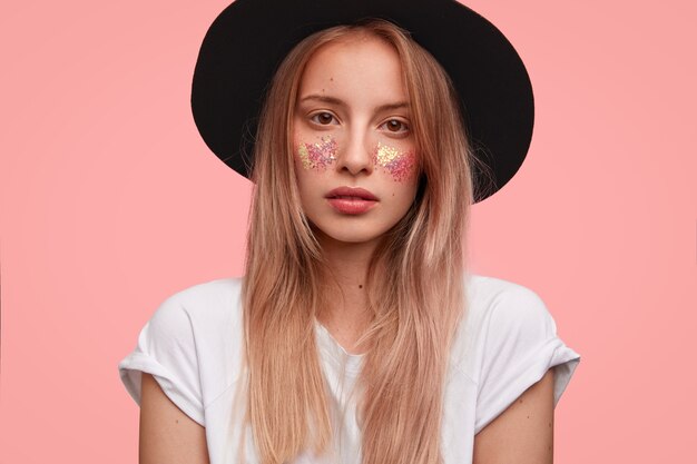 Portrait of young blonde woman with glitter on face and stylish hat