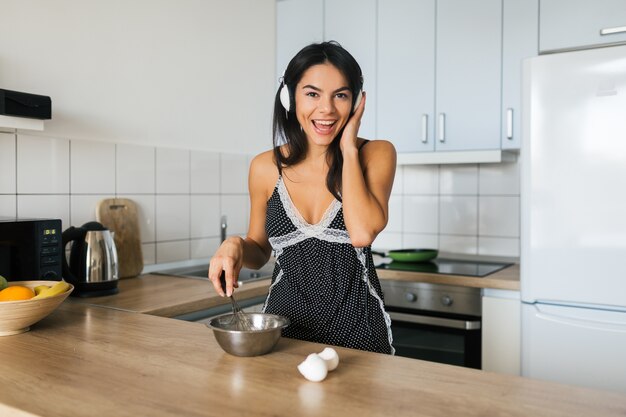 Portrait of young brunette attractive woman cooking scrambled eggs in kitchen in morning, smiling, happy mood, positive housewife, healthy lifestyle, listening to music on headphones