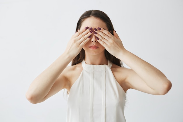 Portrait of young brunette girl covering eyes with hands .