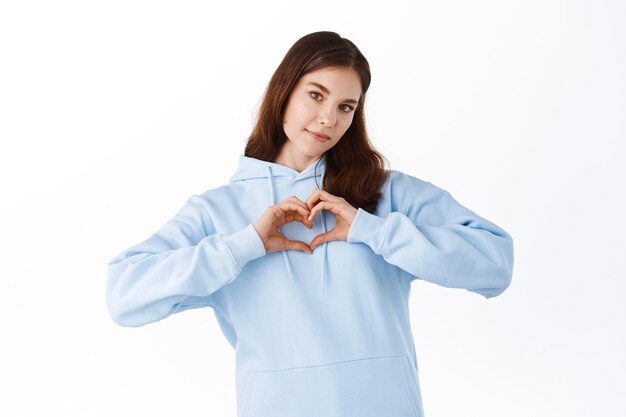 Portrait of young, brunette, pretty girl in hoodie showing with her finger heart figure, sending love to her lover, standing over white wall