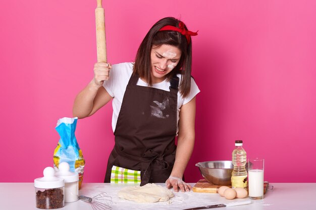 Portrait of young brunette stressed female working in kitchen whole day, preparing homemade pastry, looks tired. beats on dough with wooden rolling pin with anger isolated on rose.