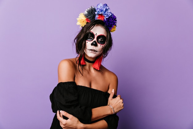 Portrait of young brunette with multi-colored crown of flowers. woman in skeleton mask looks mysteriously