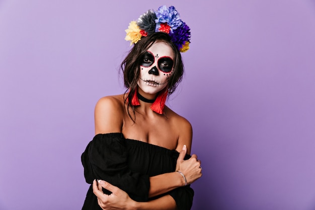 Portrait of young brunette with multi-colored crown of flowers. Woman in skeleton mask looks mysteriously
