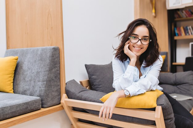 Portrait young brunette woman in black glasses chilling on couch in modern apartment. Comfortable, cheerful mood, smiling