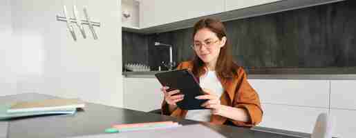 Free photo portrait of young brunette woman in glasses sitting in her kitchen with digital tablet reading on