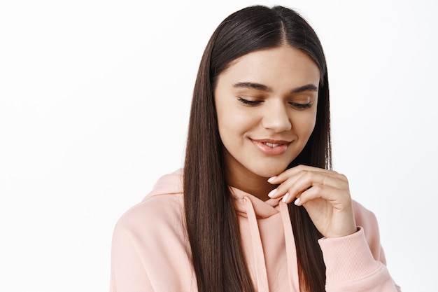 Portrait of young brunette woman hiding her gaze, looking down and smiling sensual, touching clean smooth face with natural make up, standing against white wall