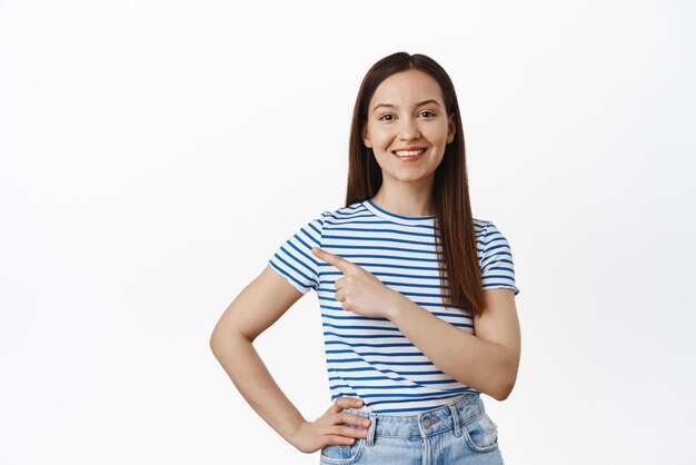 Portrait of young brunette woman pointing finger left showing sale promo point at logo smiling happy recommend click on link standing over white background
