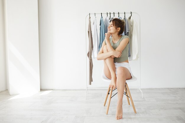 Портрет молодой женщины брюнет усмехаясь смотрящ в стороне сидя на стуле над шкафом вешалки и белой стеной.