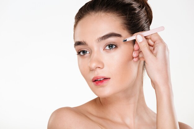 portrait of young brunette woman with soft skin plucking eyebrows with tweezers