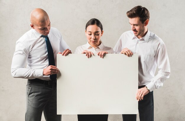 Portrait of young business people looking at big blank placard standing against grey wall