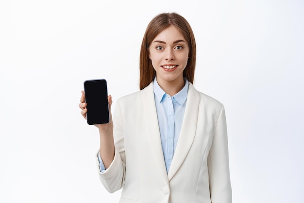 Портрет молодой деловой женщины показывает пустой мобильный экран и улыбается, демонстрируя, что финансовое приложение стоит на белом фоне