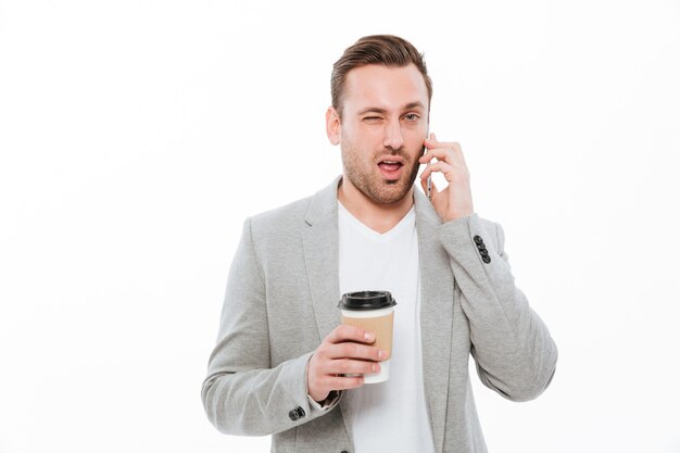 Portrait of young businessman drinking takeaway coffee while having pleasant mobile conversation on cellphone and winking, over white wall