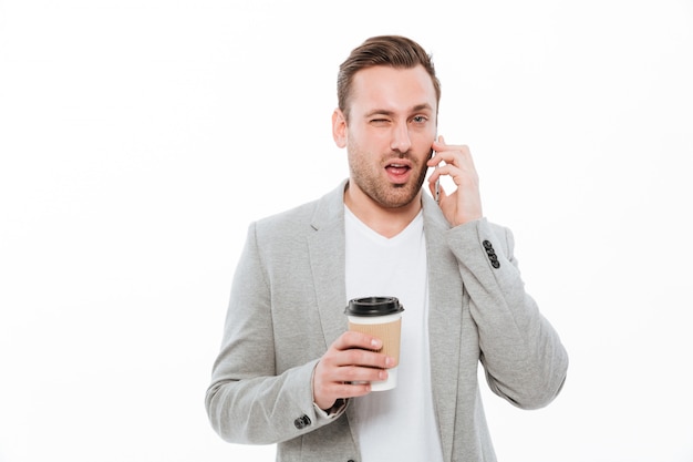 Портрет молодого бизнесмена, пьющего кофе на вынос, приятного мобильного разговора по мобильному телефону и подмигивающего над белой стеной