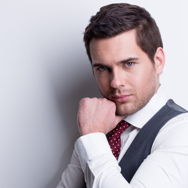 Portrait of young businessman in gray suit with hand near face poses over wall.