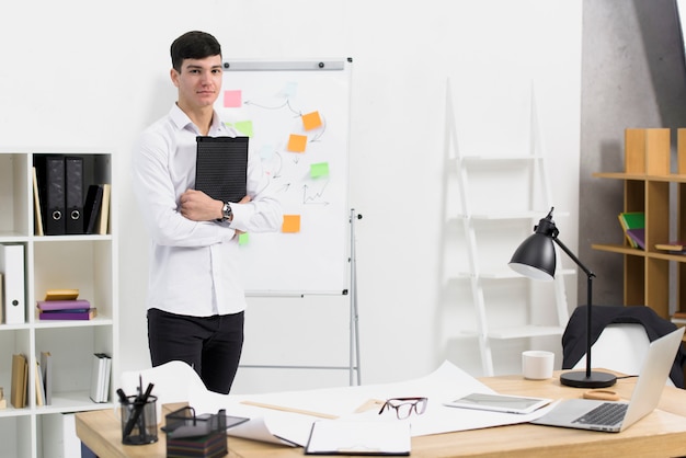 Portrait of a young businessman holding clipboard in hand standing at workplace in the office