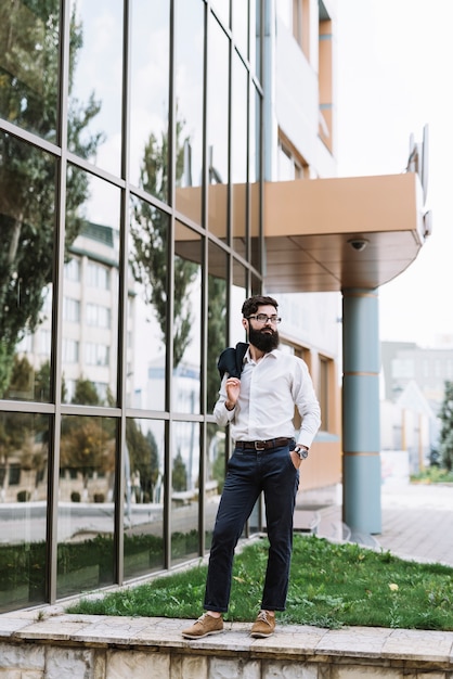 Portrait of young businessman holding coat over his shoulder standing near the corporate building