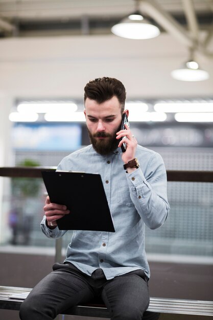 Portrait of a young businessman looking at clipboard talking on mobile phone