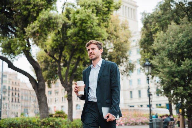 Портрет молодого бизнесмена стоя перед зданием держа кофейную чашку на вынос и цифровую таблетку