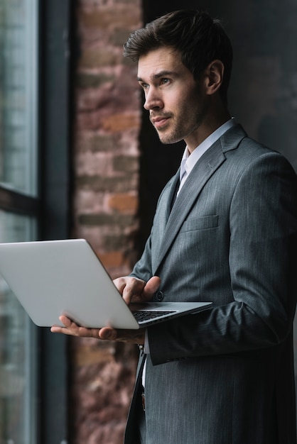 Free photo portrait of young businessman standing near the window using laptop