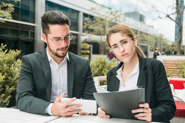 Portrait of a young businesswoman holding folder in hand sitting near the businessman looking at mobile phone