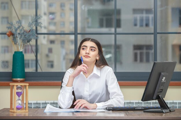 Портрет молодой деловой женщины, сидящей за столом и подносящей ручку к подбородку, думая Фото высокого качества