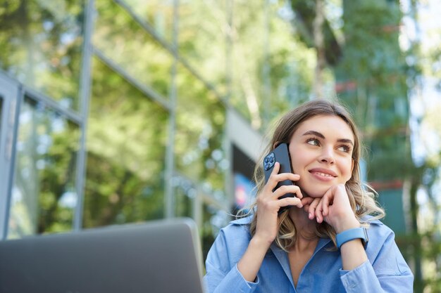 Портрет молодой деловой женщины, сидящей снаружи в парке и разговаривающей по мобильному телефону с помощью ноутбука