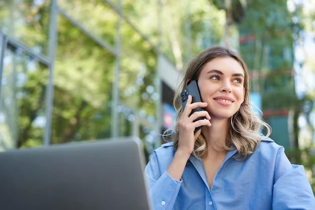 Free photo portrait of young businesswoman sitting outside in park talking on mobile phone using laptop young w