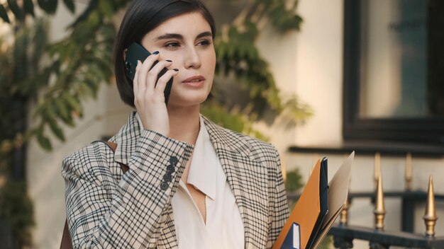 Портрет молодой деловой женщины, разговаривающей по телефону в ожидании деловой встречи на уютной городской улице. Современный агент по недвижимости ждет клиентов на открытом воздухе