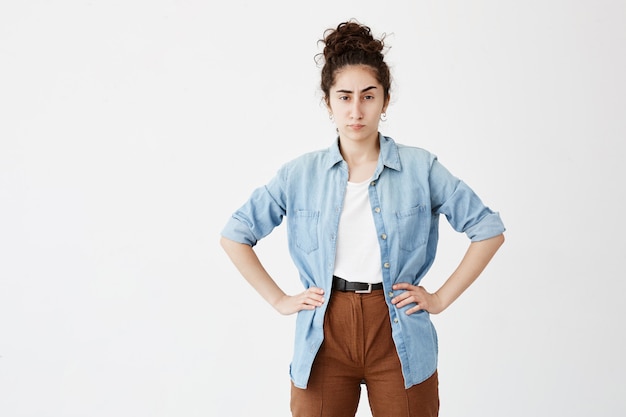 Portrait of young businesswoman with dark hair in bun wearing casual clothes, looking with annoyance, standing with hands on her hips, angry with her employees showing unsatisfactory results of work