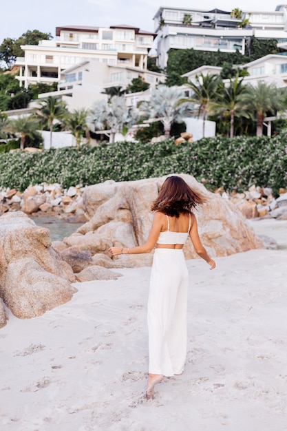 portrait of young calm happy caucasian fit slim woman in crop cami top and pants set alone on rocky tropical beach at sunset