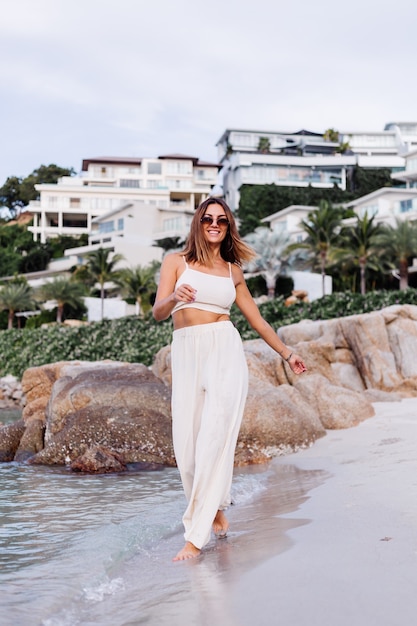 portrait of young calm happy caucasian fit slim woman in crop cami top and pants set alone on rocky tropical beach at sunset