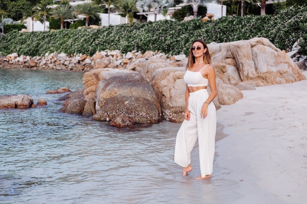 portrait of young calm happy caucasian fit slim woman in crop cami top and pants set alone on rocky tropical beach at sunset