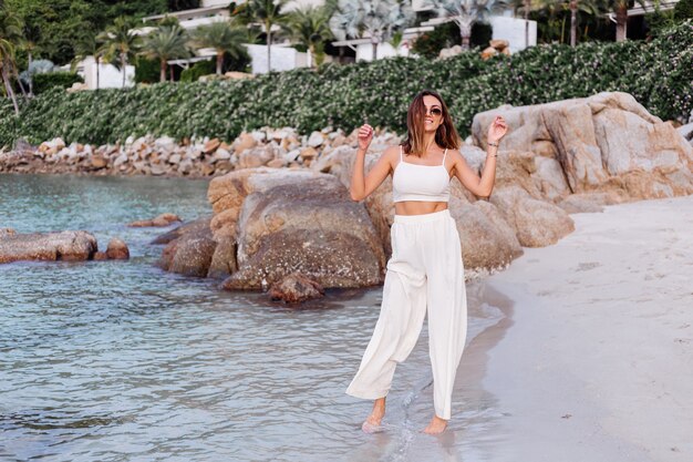 portrait of young calm happy caucasian fit slim woman in crop cami top and pants set alone on rocky tropical beach at sunset